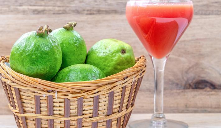 Fotografia de um refresco de leite ninho, neston e suco de goiaba em uma taça de vidro, e ao lado tem uma cesta cheia de goiabas que está sobre uma mesa de madeira clara.
