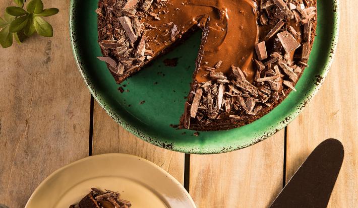 Fotografia em tons de verde em uma bancada de madeira clara com um suporte verde com o bolo de chocolate em cima. Ao lado, um prato com uma fatia do bolo e uma espátula para bolo.