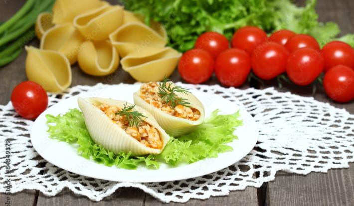 Fotografia de duas unidades de macarrão tipo concha recheado com creme de palmito sobre uma folha de alface, que está em um prato redondo branco e pequeno. Ao fundo, algumas unidades de macarrão tipo concha crus e ao lado, tomates cereja.