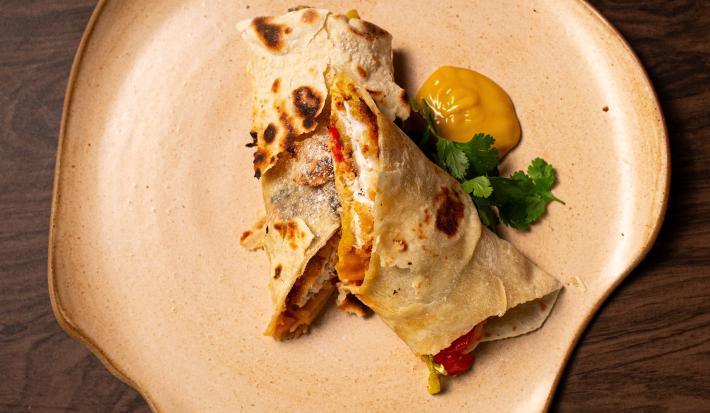 Fotografia de um wrap de moqueca cortado na metade, e cada metade sobre a outra. Ao lado do wrap tem um molho de mostarda e salsinha.