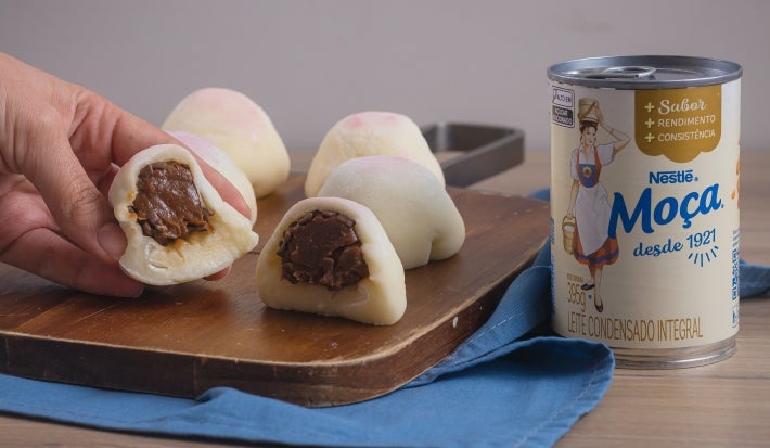 Receita de Mochi de Brigadeiro, servida em pequenas bolinhas brancas, recheadas com brigadeiro enrolado, sobre uma tábua de madeira e um tecido na cor azul ao lado de uma Lata de Leite Moça
