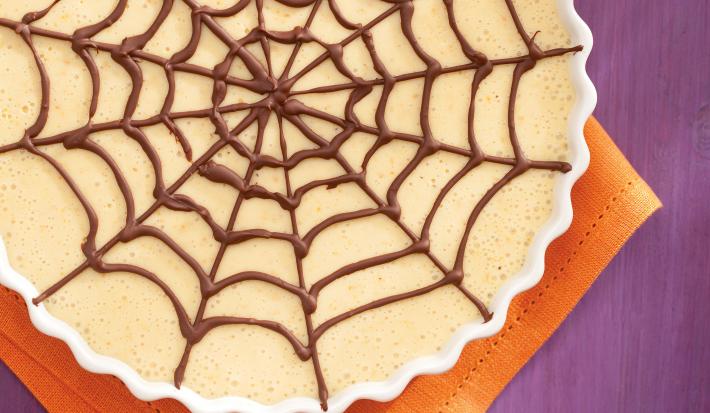 Fotografia em tons de roxo, laranja e marrom de uma bancada roxa vista de cima, um pano laranja e por cima um recipiente branco e redondo com a sobremesa de gelatina e para decorar chocolate por cima em forma de teia de aranha