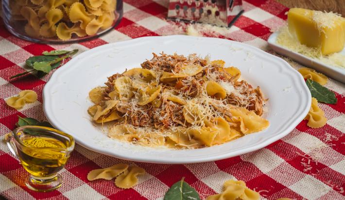 Foto de uma mesa com uma toalha xadrez vermelha e branco. Sobre ela já macarrões crus, um pedaço de queijo, um ralador, um pote com azeite e algumas folhas. No centro há um prato branco com a receita de Farfalle com Ragú de Frango.