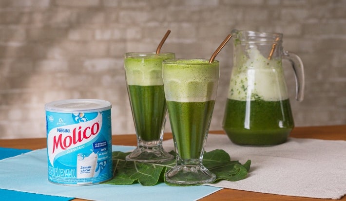 Fotografia em tons de verde com dois copos grandes ao centro. Dentro dos copos existe uma receita de suco verde feita com couve e Molico.