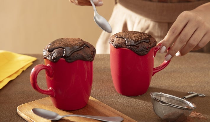 Fotografia de duas canecas vermelhas com bolo e cobertura de nescau extra cacau, ao lado de uma colher de sopa e uma mini peneira. A foto mostra uma mão femnina segurando uma das canecas.