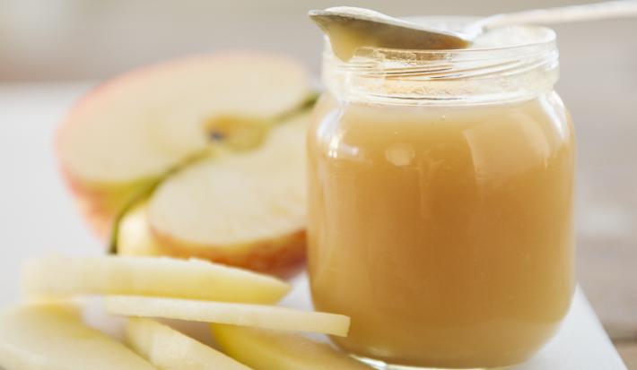 Fotografia de um potinho de vidro com um creme de maçã com iogurte e farofa doce. Apoiada no potinho, uma colher de sobremesa, e ao lado, cinco fatias de maçã sem casca junto com uma metade da maçã cortada ao meio, sobre uma tábua de cor branca.