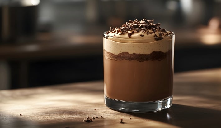 Fotografia da sobremesa com o creme com doce de leite e chocolate, kitkat e raspas de chocolate por cima, dentro de um copo de vidro pequeno, sobre uma mesa de madeira.