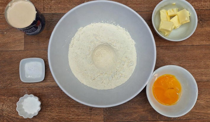 Farinha com uma cova no centro dentro de uma tigela ao centro da imagem. Tigelas dispostas ao redor, cada uma com um ingrediente: manteiga, ovos, fermento e sal. Uma lata aberta de creme de leite Nestlé