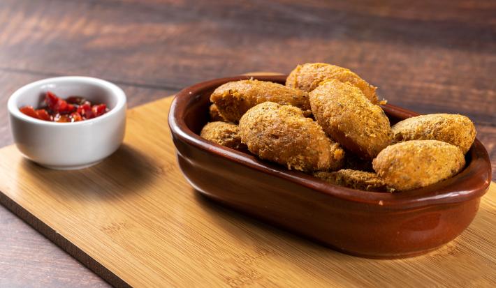 Fotografia de uma travessa marrom com vários bolinhos fritos em tons dourados por cima de uma tábua de madeira. Ao lado, um pequeno recipiente branco com um molho de pimentas vermelho.