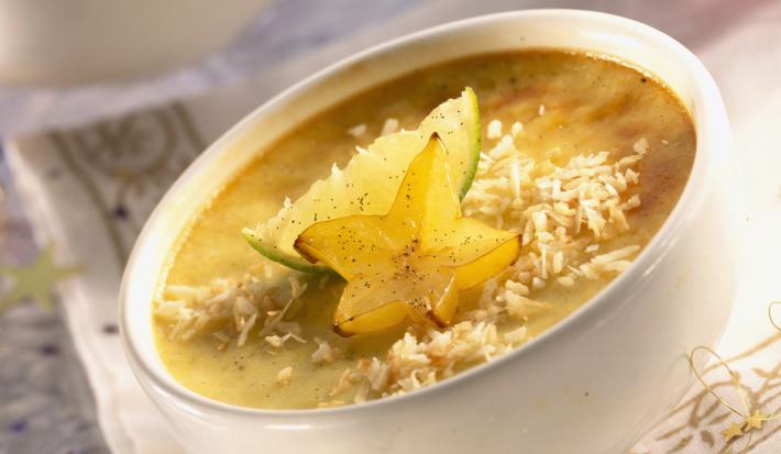 Fotografia de um flan de carambola com coco ralado por cima, limão e uma rodela de carambola. O creme está dentro de um recipiente pequeno de vidro na cor branco, que está sobre uma toalha de mesa branca com detalhes dourados.