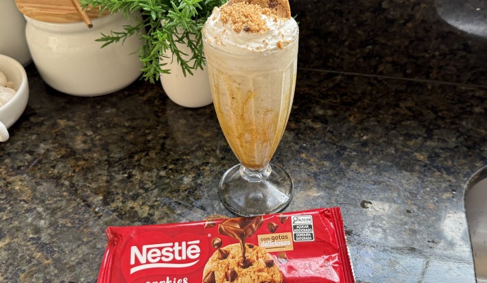 Foto da receita de Milkshake de Cookies e Caramelo, servido em um copo alto, numa bancada de mármore com um pacote de Cookies Nestlé Tradicional à frente