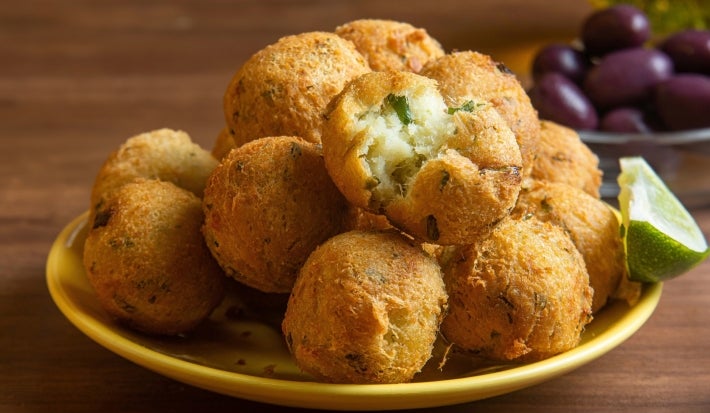 Em cima de uma mesa, prato amarelo com porção de bolinhos de bacalhau dourados e crocantes. No lado direito, há um bolinho aberto ao meio com bacalhau cremoso.