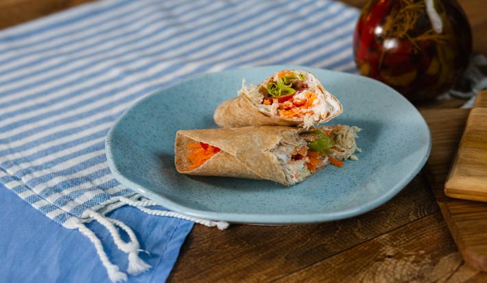 Fotografia em tons de azul com um prato azul ao centro. Em cima do prato existe uma receite de wrap recheada com frango desfiado, creme de ricota, tomate e alface.