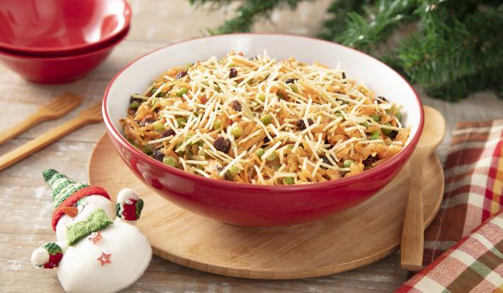 Foto da receita de Salpicão de Natal Vegano. Observa-se um recipiente refratário vermelho de porcelana com o salpicão dentro, coberto de batata palha. Observa-se decorações natalinas enfeitando a foto, como galhos de pinheiro e um papai noel à frente.