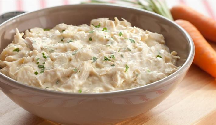 Em uma bancada de madeira cor bege contém um recipiente redondo branco com pedaços de frango e creme branco. Ao lado duas cenouras