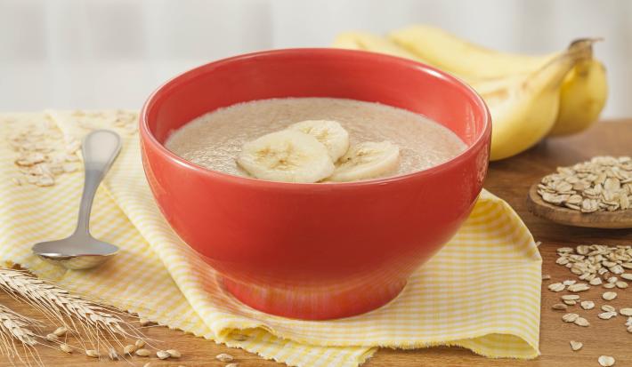 Fotografia em tons de amarelo e vermelho de uma bancada de madeira com um paninho amarelo e um recipiente redondo vermelho, sobre ele o mingau de farinha láctea com fatias de banana. Ao fundo duas bananas e aveias espalhadas pela bancada.