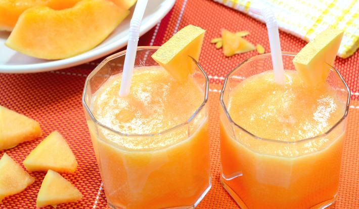 Fotografia de dois copos de vidro com vitamina de melão e maçã, com um pedaço de melão decorando do copo. Ao lado, pedaços de melão sobre uma toalha de mesa vermelha, e no fundo, um prato branco com fatias de melão.