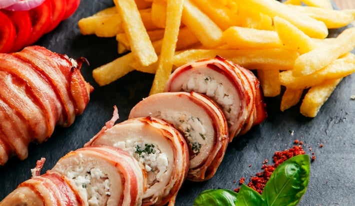 Fotografia em tons claros de dois tornedores de frango recheados enrolados no bacon, ao redor tem tomates, cebola roxa fatiada, batatas fritas e algumas folhas, sobre um apoio quadrado preto. O apoio está sobre uma mesa de madeira.