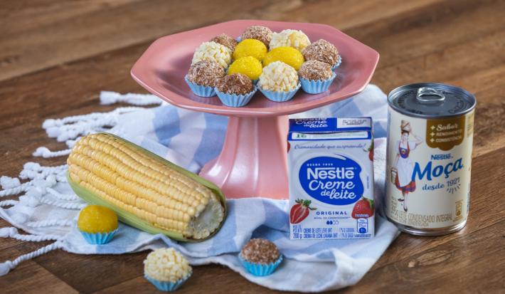 Foto em tons bem coloridos da receita de brigadeiro de milho em várias porções servidas sobre uma base alta cor de rosa com um paninho azul embaixo. Ao lado, há uma espiga de milho, uma caixinha de creme de leite e uma lata de leite moça