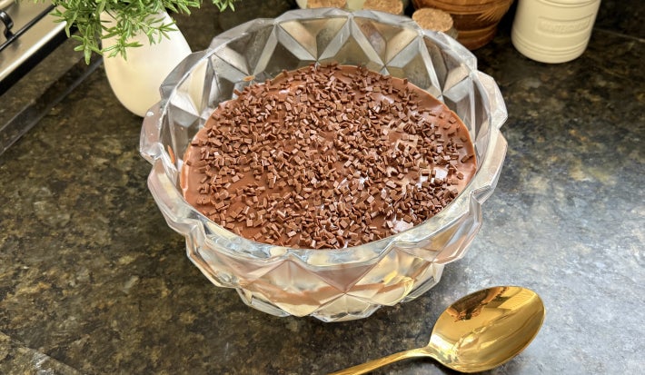 Fotografia mostra um recipiente de vidro redondo com uma cobertura de chocolate e granulado por cima sobre uma bancada preta.