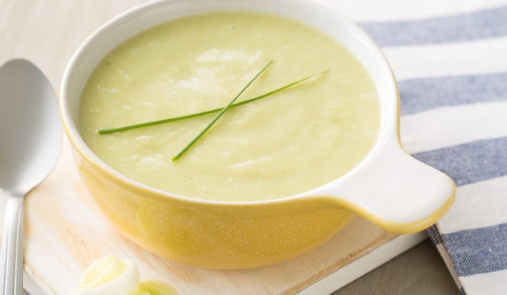 A imagem apresenta um prato de sopa cremosa de alho-poró, servida em uma tigela amarela com alça. A sopa é decorada com dois talos de cebolinha. Ao lado, há fatias de alho-poró. Uma colher prateada está à esquerda, e um pano listrado azul e branco