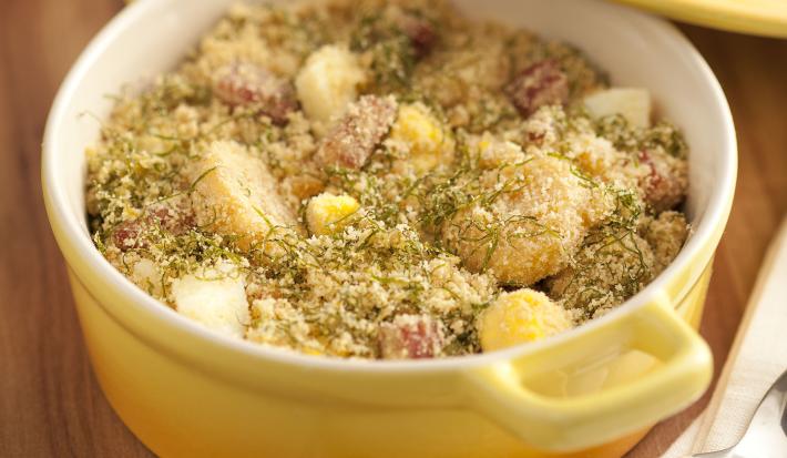 Fotografia em tons de amarelo em uma bancada de madeira escura com uma panela de cerâmica amarela com alças laterais e a farofa de banana com couve dentro. Ao lado, a tampa da panela e guardanapo de pano branco.