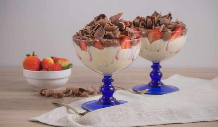 Foto da receita de pavê de leite moça com morangos e chocolate, servidos em taças com uma base azul, sobre uma bancada de madeira decorada com um tecido, pedaços de chocolate, um pote com morangos e uma lata de Leite Moça