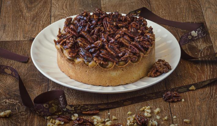 Foto da receita de Torta de Caramelo Salgado e Castanhas Caramelizadas, servida num prato redondo branco, sobre uma bancada de madeira, decorada com uma fita de páscoa e castanhas picadas