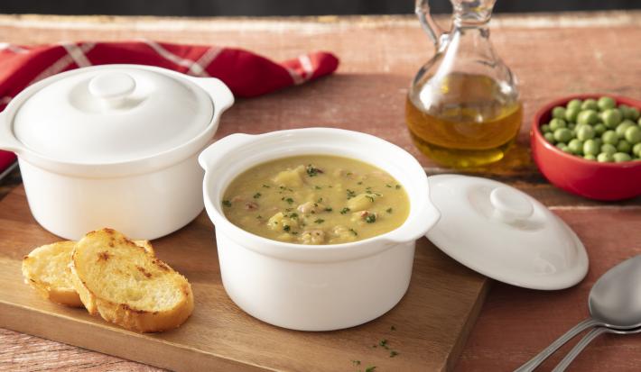 Fotografia em tons de marrom e vermelho de uma bancada de madeira, ao centro uma tábua com 2 mini caçarolas brancas, em uma delas, é possível ver o creme verde da sopa de ervilha. Ao fundo, azeite e ervilhas congeladas.