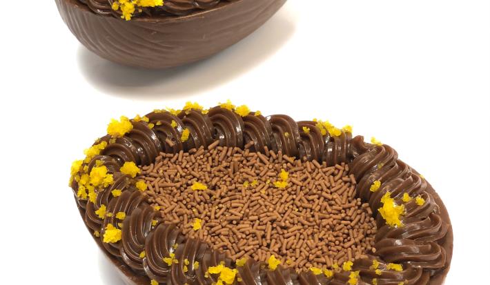 Foto em tons de branco, marrom claro, escuro e laranja. Na foto é possível ver uma bancada branca com duas cascas de ovos recheadas com brigadeiro, chocolate granulado e migalhas de bolo de cenoura