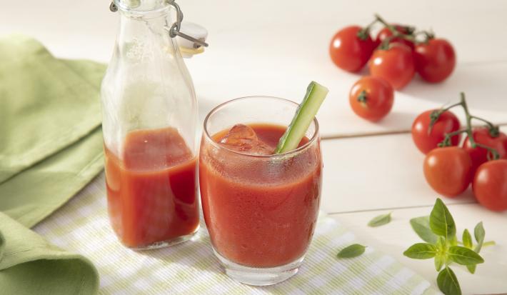 Fotografia em tons de vermelho em uma bancada de madeira clara, um pano verde liso e outro xadrez, um copo de vidro com o suco de tomate e uma jarra com mais suco ao centro da foto. Ao lado, tomates pequenos para decorar a mesa.