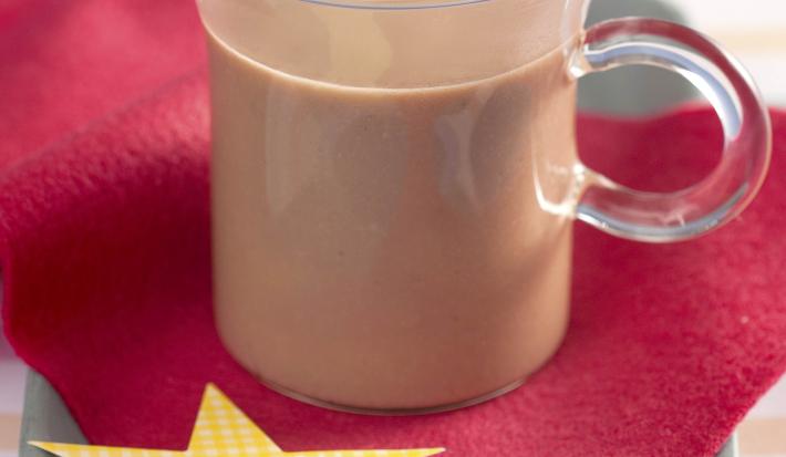 Fotografia em tons de vermelho e azul em uma bancada de madeira azul, uma toalhinha listrada em azul e rosa, um paninho vermelho com uma estrela amarela. Ao centro, uma xícara de vidro com chocolate quente.