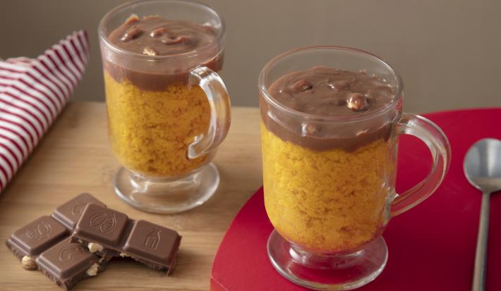 Fotografia em tons de laranja em uma bancada de madeira de cor marrom. Ao centro, uma tábua de madeira vermelha contendo duas taças de vidro com os bolos. Ao lado, há uma colher de inox e alguns tabletes de chocolate. Ao fundo, um pano branco listrado.
