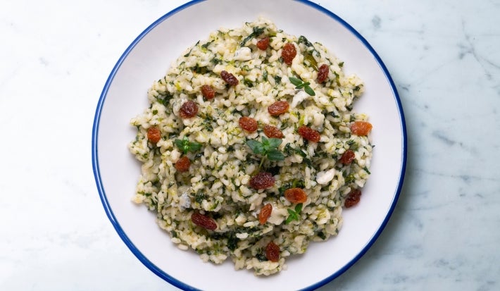 Visto de cima, prato redondo branco com porção de arroz cremoso verde e uvas-passas. Decorado com uma folha de manjericão.