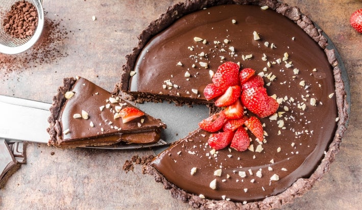 Fotografia vista de cima de uma torta grande com base de chocolate, recheio de creme branco e ganache por cima, decorada com morangos e castanhas trituradas. A foto mostra uma faca grande segurando uma fatia da torta.