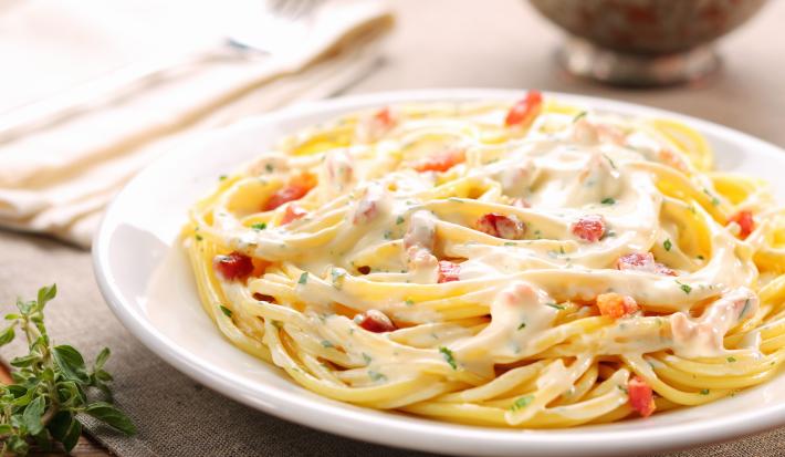 Fotografia de um prato de macarrão cozido, coberto com molho branco cremoso e bacon crocante.