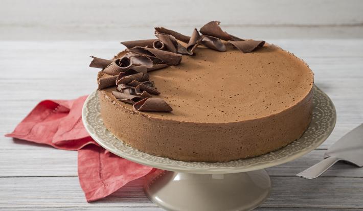 Fotografia em tons de branco e vermelho de uma bancada de madeira branca com um paninho vermelho, sobre ele uma bailarina branca com a torta mousse de chocolate decorada com raspas de chocolate.