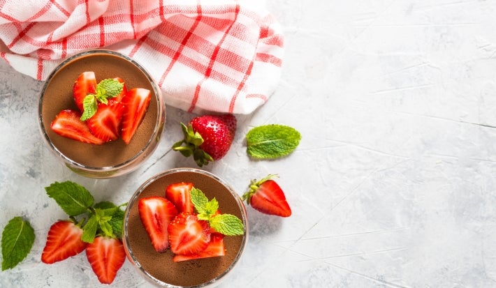 Fotografia em tons claros e vista de ciam de dois copos decorativos de tamanho normal com a sobremesa de taça moranguda, finalizada com chocolate e decoração de morangos em pedaços e folhas de hortelã.
