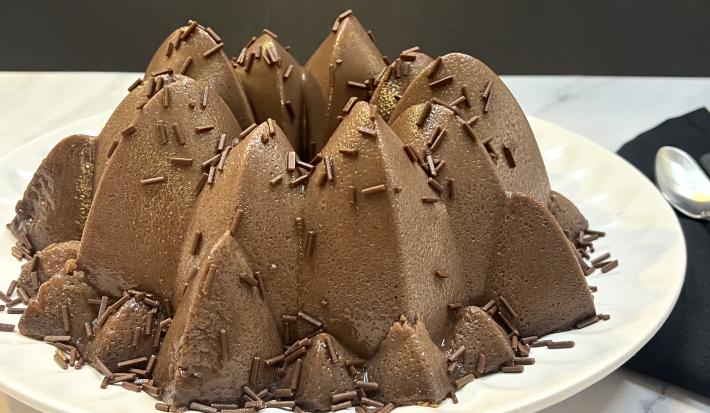 Foto aproximada da receita de Brigadeirão Tradicional, feito em uma forma com formato diferente, servido num prato redondo branco e decorado com granulados.