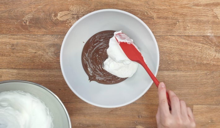 Mão misturando claras em neve com ganache de chocolate com espátula vermelha.