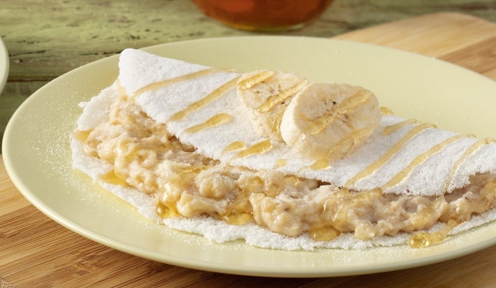 Fotografia de uma um prato redondo de tom claro, sobre ele uma tapioca. O prato está apoiado em uma tábua de madeira.