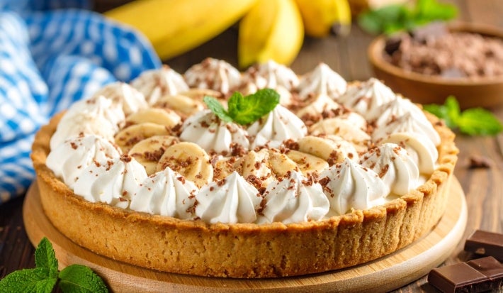 Em cima de uma mesa, banoffe com massa crocante, decorado com chantilly cremoso, bananas cortadas em rodelas e chocolate ralado.