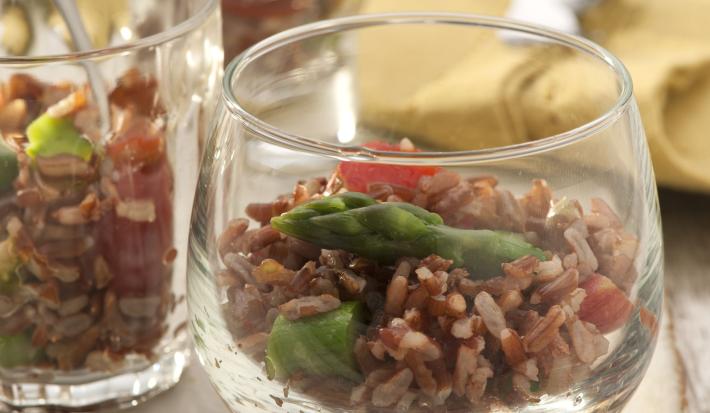 Fotografia em tons de marrom de uma bancada de madeira com uma tábua quadrada marrom, sobre ela um recipiente de vidro com salada de arroz vermelho. Ao fundo um paninho amarelo.