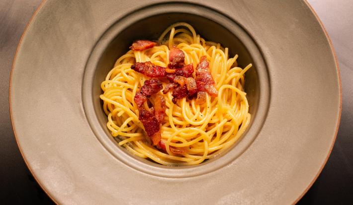Fotografia em tons de amarelo com um prato cinza ao centro. Dentro do prato existe uma receita de macarrão carbonara finalizada com pedaços de guanciale frito.
