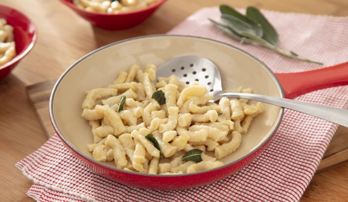 Foto da receita de Spatzle. Observa-se uma frigideira grande sobre um pano de prato quadriculado vermelho. A massa está dentro da frigideira com folhas de manjericão.