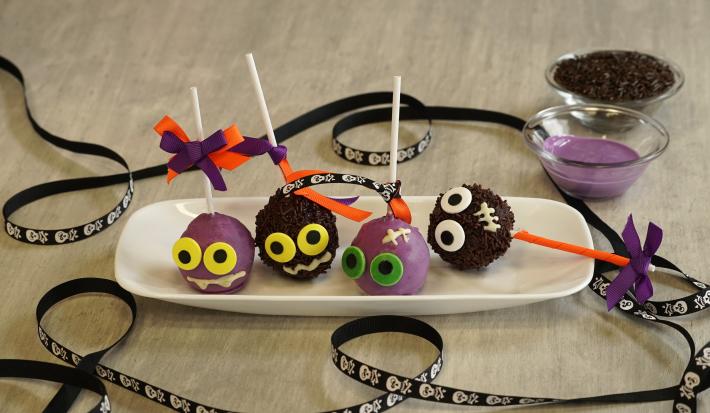 Imagem de um prato branco com 4 bolinhas decoradas com palitos brancos. A cena está decorada com uma fita preta com caveiras brancas e dois potes com chocolate roxo e granulados pretos.