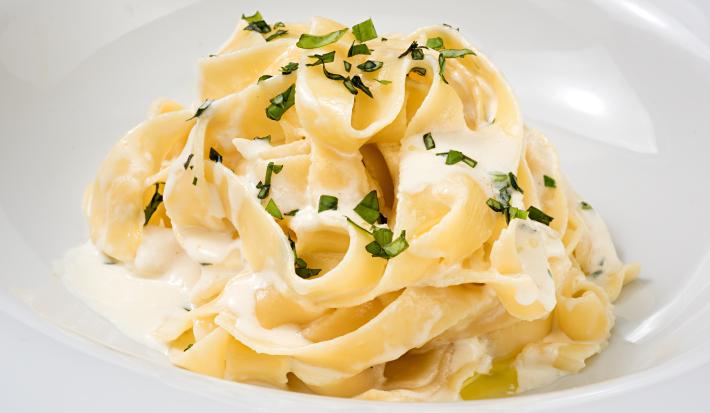 fotografia em tons de branco e amarelo tirada de um prato redondo branco, que contém um macarrão com requeijão e manjericão por cima.