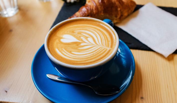 Foto da receita de Flat White. Observa-se uma xícara azul sobre um pires da mesma cor e a bebida dentro, com um formato de coração.