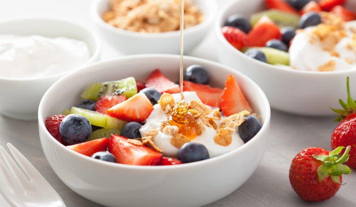 Fotografia em tons de branco, com três refratários brancos fundos no canto e no centro outro refratário com diversas frutas, iogurte e uma calda de mel caindo. No canto inferior direito, dois morangos inteiros.