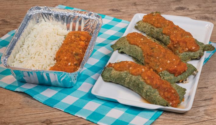 Foto de uma marmita de alumínio com arroz e panqueca verde dentro. Ao lado há um prato branco retangular com três panquecas verdes cobertas com molho de tomate. Tudo está sobre um pano azul e branco quadriculado numa mesa de madeira
